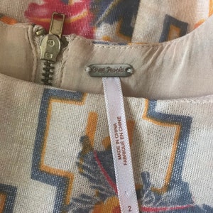 May include: Close-up of a cream-colored dress with a floral and geometric pattern in shades of red, yellow, and blue. The dress features a metal zipper and a tag that reads "Free People" and "Made in China". Size 2.