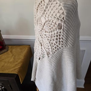 May include: A cream-coloured, open-weave crochet sweater with a unique design on the back. The sweater features a ribbed knit pattern on the front and sleeves. The back has a large, open crochet pattern. The sweater is displayed on a mannequin.