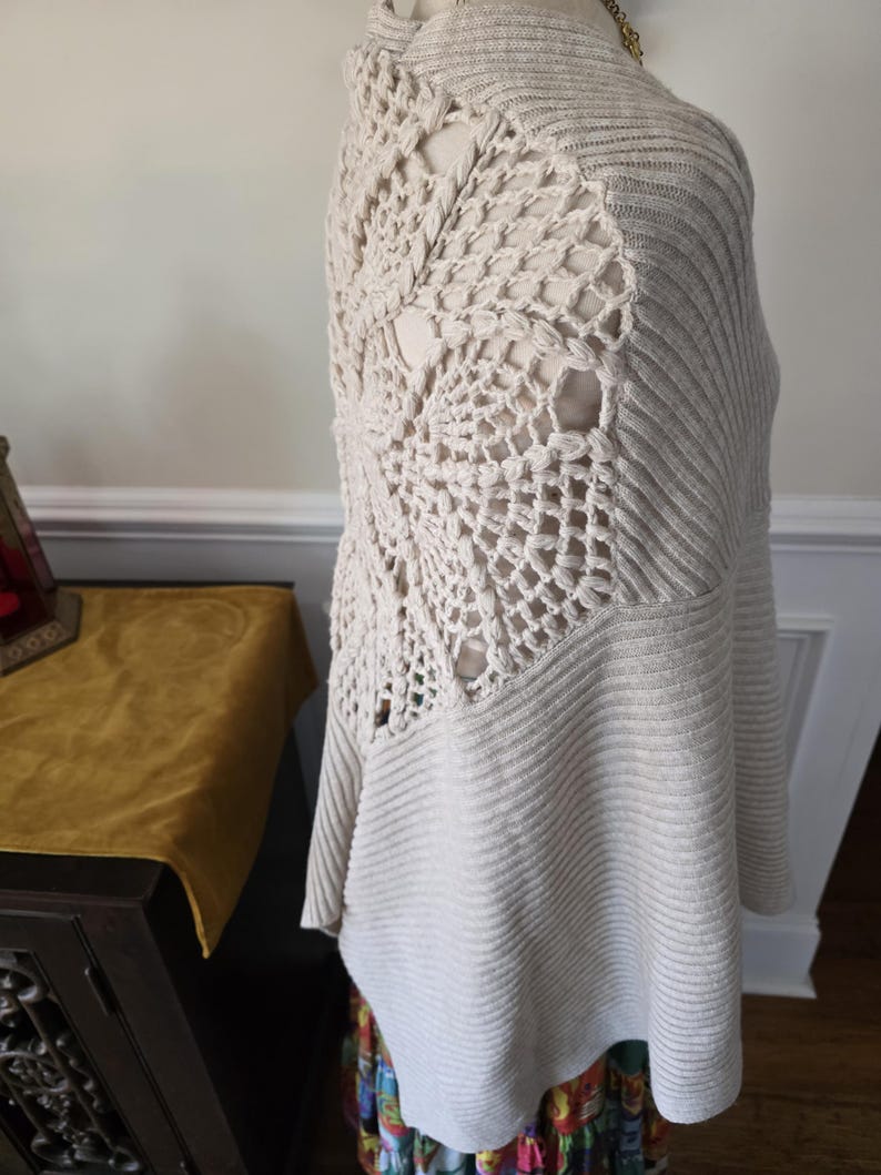 May include: A cream-coloured, open-weave crochet sweater with a unique design on the back. The sweater features a ribbed knit pattern on the front and sleeves. The back has a large, open crochet pattern. The sweater is displayed on a mannequin.