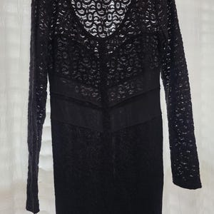 May include: Black lace dress with long sleeves and a V-neckline. The dress features a fitted bodice and a straight skirt. The lace pattern is intricate and detailed, creating a delicate and elegant look. The dress is suitable for special occasions.
