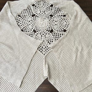 May include: A light beige knitted poncho featuring a cream-coloured, openwork crochet centre. The poncho has a ribbed texture and a draped design. The crochet detail is circular with a floral pattern. The garment is laid out on a dark brown wooden surface.