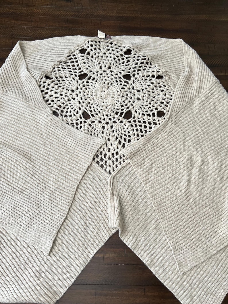 May include: A light beige knitted poncho featuring a cream-coloured, openwork crochet centre. The poncho has a ribbed texture and a draped design. The crochet detail is circular with a floral pattern. The garment is laid out on a dark brown wooden surface.