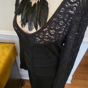 May include: A black lace dress with long sleeves and a V-neckline. The dress features a fitted bodice and a decorative black feather necklace. The dress is displayed on a mannequin.