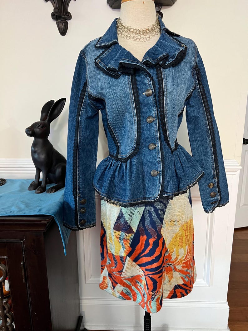 Puede incluir: Una chaqueta vaquera azul con ribete de encaje negro y un dobladillo con volantes. La chaqueta est&aacute; abotonada y tiene una silueta ajustada. La chaqueta se lleva sobre una falda estampada de colores.