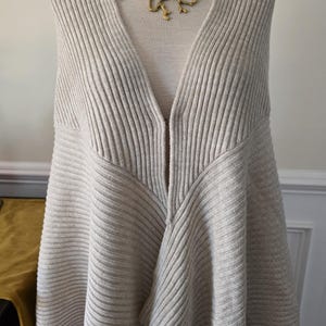 May include: A light beige, ribbed knit cape-style sweater with a deep V-neck. The sweater is displayed on a mannequin and paired with a gold-coloured, organic-shaped necklace. The knitwear has a flowing, asymmetrical design.