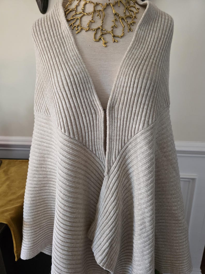 May include: A light beige, ribbed knit cape-style sweater with a deep V-neck. The sweater is displayed on a mannequin and paired with a gold-coloured, organic-shaped necklace. The knitwear has a flowing, asymmetrical design.