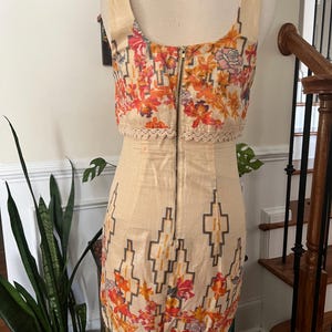 May include: A beige sleeveless dress with a floral and geometric pattern. The top section features a colorful floral print with orange, pink, and yellow flowers. The dress has a zipper down the back and a decorative lace trim.