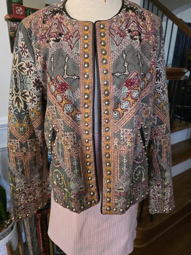 May include: A gray jacket with an intricate paisley pattern in shades of red, yellow, and green. The jacket has a black trim and is adorned with silver studs along the edges and pockets. The jacket is open in the front.