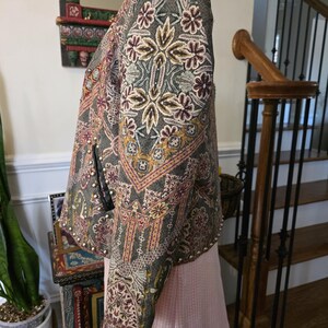 May include: A gray jacket with intricate floral embroidery in shades of cream, burgundy, and gold. The jacket features a detailed pattern and is adorned with small, silver-colored studs along the edges. The sleeves are long and the jacket is displayed on a mannequin.