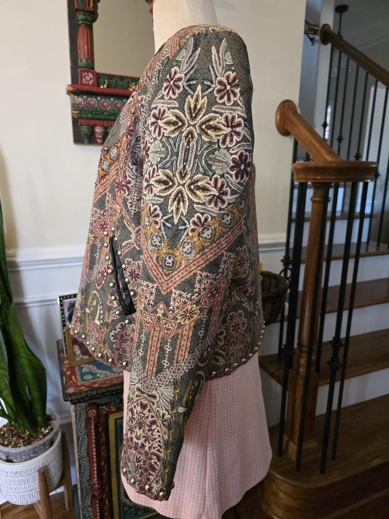 May include: A gray jacket with intricate floral embroidery in shades of cream, burgundy, and gold. The jacket features a detailed pattern and is adorned with small, silver-colored studs along the edges. The sleeves are long and the jacket is displayed on a mannequin.