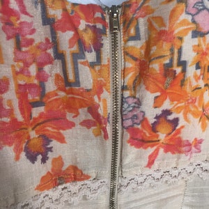 May include: Close-up of a cream-colored garment with a floral pattern in shades of orange, pink, and purple. The garment features a gold zipper down the center and delicate white lace trim along the bottom edge. The fabric has a subtle shimmer.