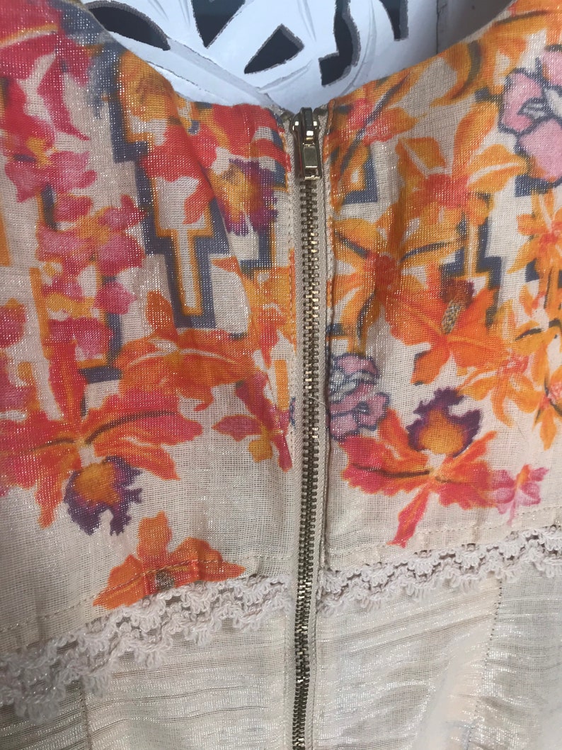 May include: Close-up of a cream-colored garment with a floral pattern in shades of orange, pink, and purple. The garment features a gold zipper down the center and delicate white lace trim along the bottom edge. The fabric has a subtle shimmer.