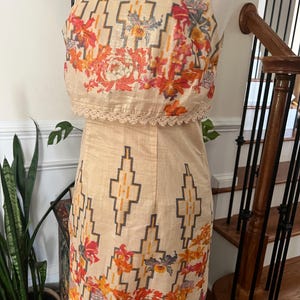 May include: A two-piece beige dress with a floral and geometric pattern. The top is a crop top with a lace trim, and the skirt is a pencil skirt. The dress has a scoop neckline and is sleeveless. A peach-colored necklace is also visible.