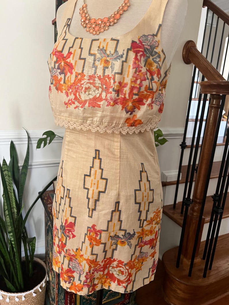 May include: A two-piece beige dress with a floral and geometric pattern. The top is a crop top with a lace trim, and the skirt is a pencil skirt. The dress has a scoop neckline and is sleeveless. A peach-colored necklace is also visible.