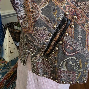May include: A close-up of a patterned jacket with gold-colored studs. The jacket features a paisley design in shades of gray, orange, and purple. Black leather trim accents the front. The jacket is paired with a light pink garment.