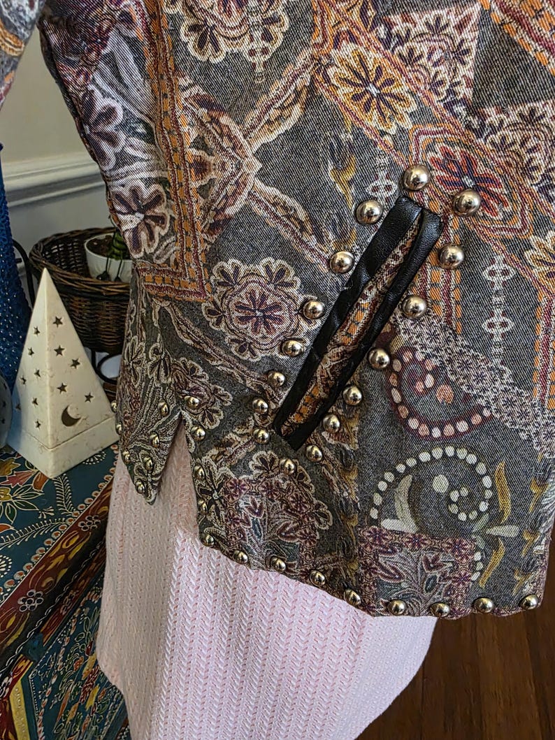 May include: A close-up of a patterned jacket with gold-colored studs. The jacket features a paisley design in shades of gray, orange, and purple. Black leather trim accents the front. The jacket is paired with a light pink garment.