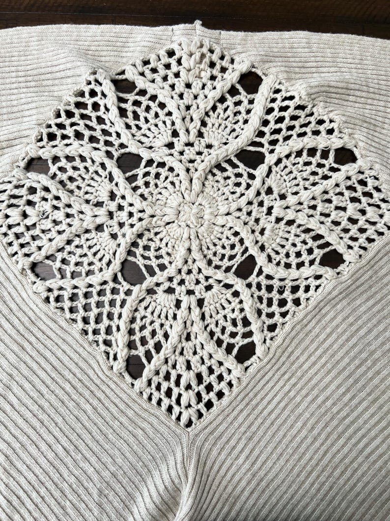 May include: A light grey sweater featuring a large, cream-coloured, crocheted square design on the front. The crocheted design has a central floral motif and intricate openwork patterns. The sweater has a ribbed texture.