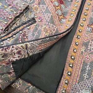 May include: Close-up of a gray jacket with an intricate paisley pattern in shades of purple, orange, and yellow. The jacket features silver-colored button details and black trim. The lining is black.