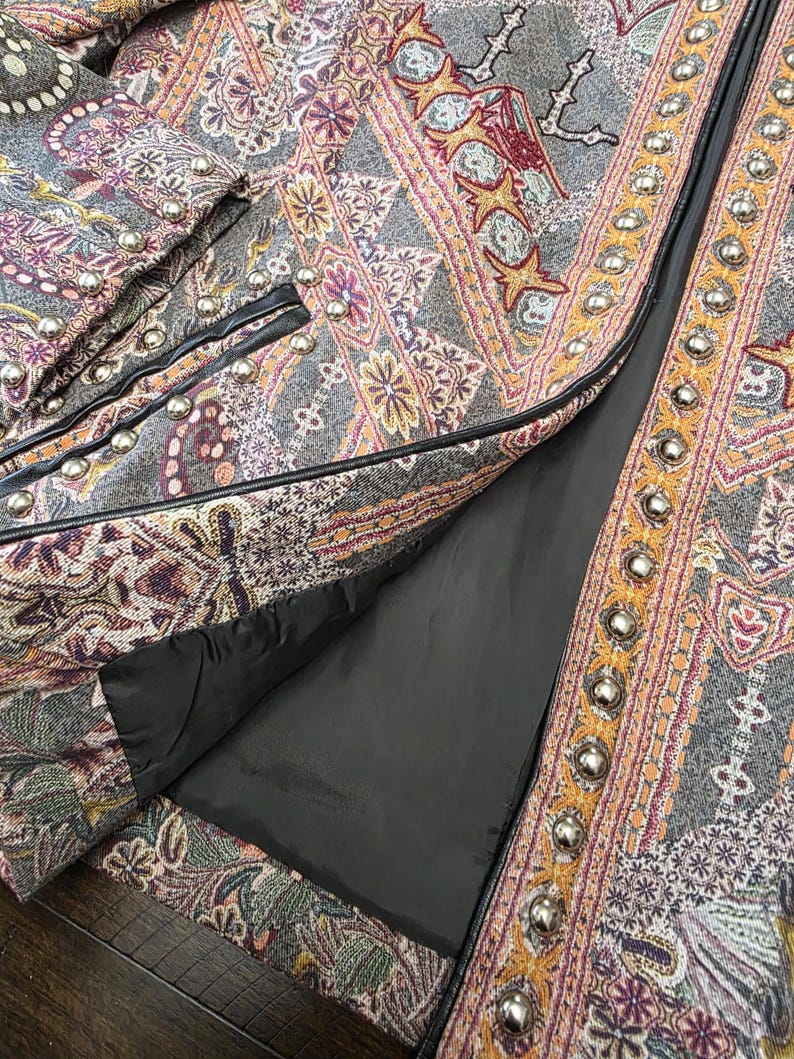 May include: Close-up of a gray jacket with an intricate paisley pattern in shades of purple, orange, and yellow. The jacket features silver-colored button details and black trim. The lining is black.