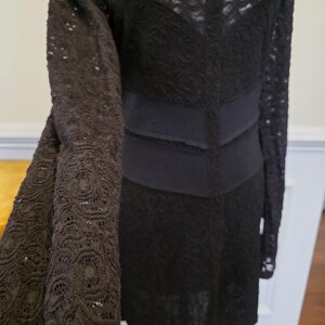 May include: Black lace dress with long sleeves. The dress features a scoop neckline and a fitted bodice with a defined waist. The sleeves and skirt are made of a delicate, openwork lace, creating a textured look. The dress is a solid black color.