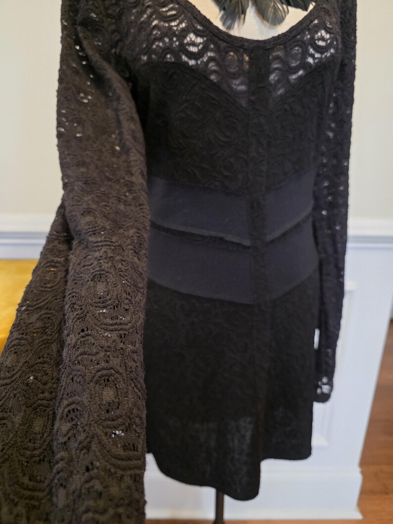 May include: Black lace dress with long sleeves. The dress features a scoop neckline and a fitted bodice with a defined waist. The sleeves and skirt are made of a delicate, openwork lace, creating a textured look. The dress is a solid black color.