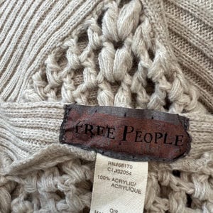 May include: Close-up of a beige knitted sweater with a textured pattern. The sweater features a label with the brand name "FREE PEOPLE" and a tag with the material composition and origin.