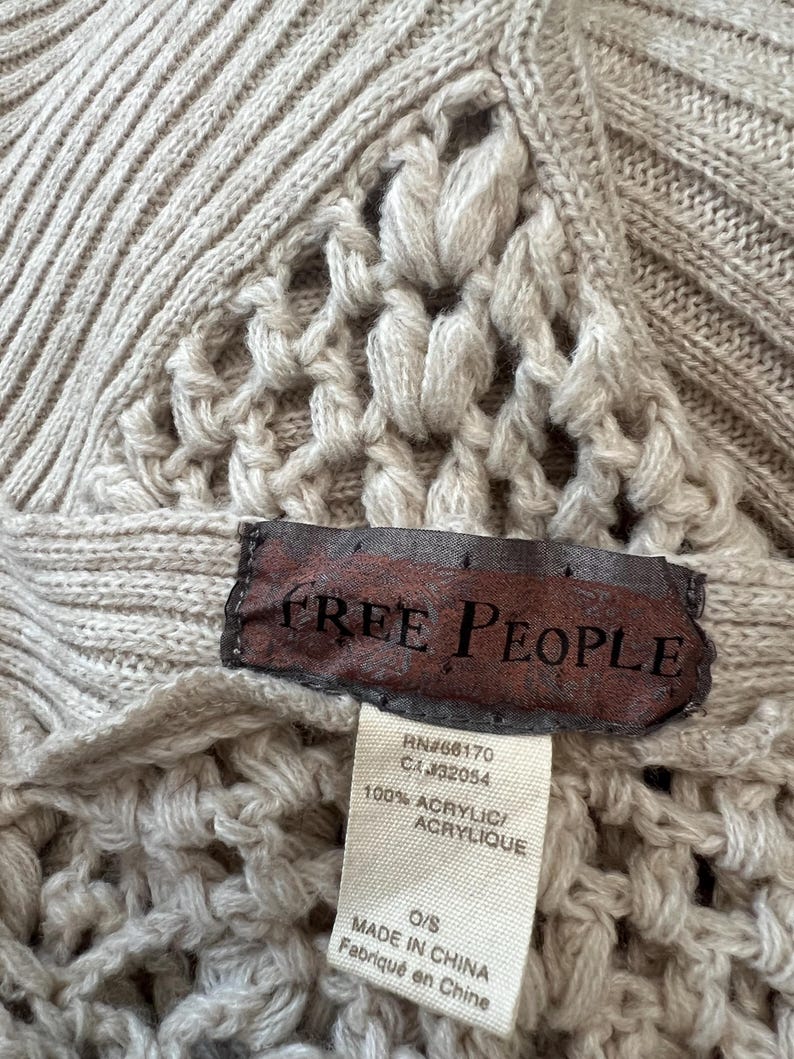 May include: Close-up of a beige knitted sweater with a textured pattern. The sweater features a label with the brand name "FREE PEOPLE" and a tag with the material composition and origin.