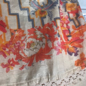 May include: Close-up of a beige fabric with a floral and geometric pattern. The design features orange, red, and yellow flowers, along with a gray and orange geometric pattern. The fabric has a metallic sheen and a white lace trim.