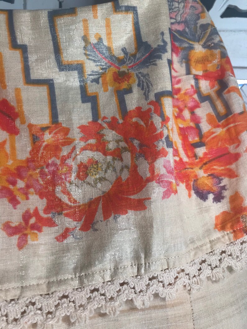 May include: Close-up of a beige fabric with a floral and geometric pattern. The design features orange, red, and yellow flowers, along with a gray and orange geometric pattern. The fabric has a metallic sheen and a white lace trim.