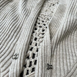 May include: A light beige cardigan with a ribbed knit design. The centre panel features a crocheted pattern. The cardigan has two metal clasps for closure. The overall style is casual and comfortable.