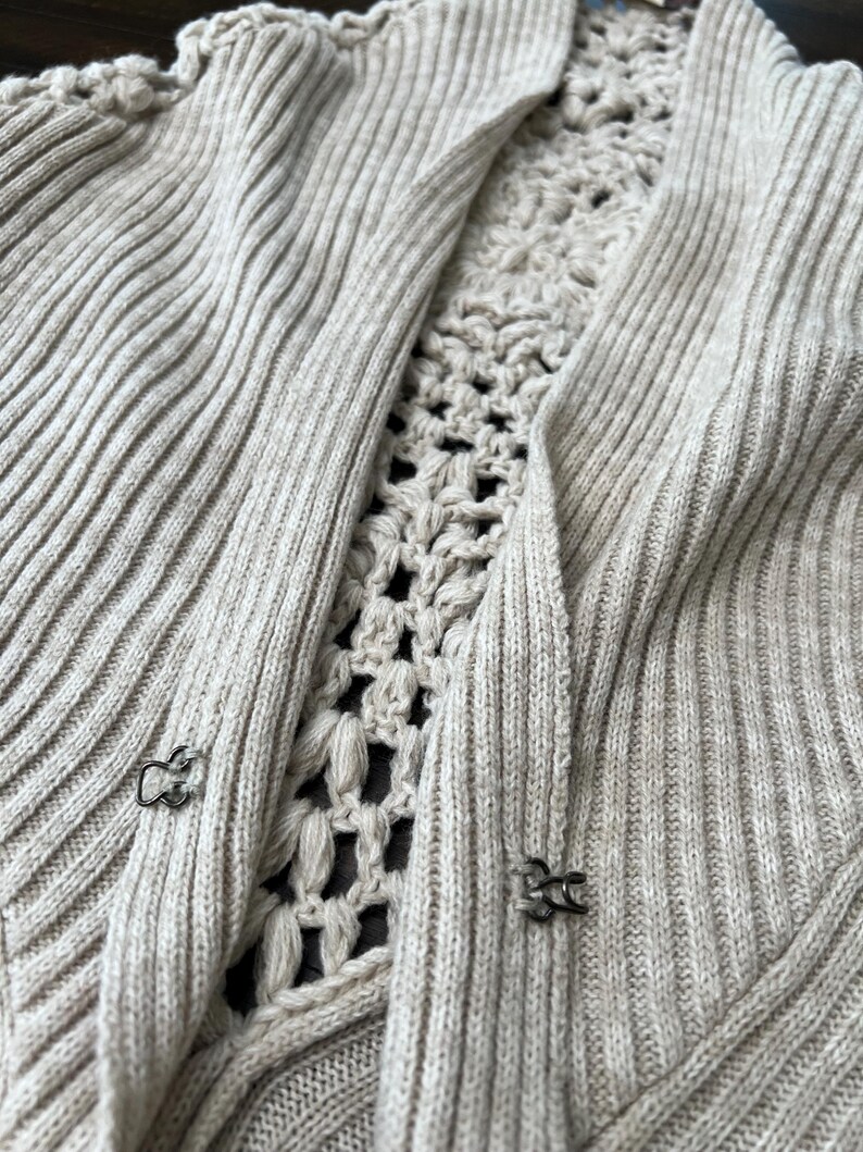 May include: A light beige cardigan with a ribbed knit design. The centre panel features a crocheted pattern. The cardigan has two metal clasps for closure. The overall style is casual and comfortable.