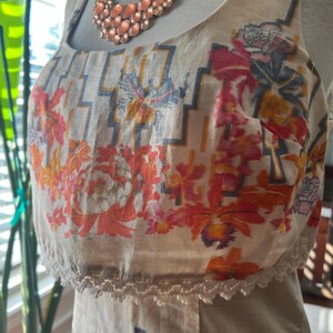 May include: A beige sundress with a floral print in shades of orange, pink, and blue. The dress features a scoop neckline, a decorative lace trim, and a geometric pattern. A peach-colored necklace is draped over the dress.