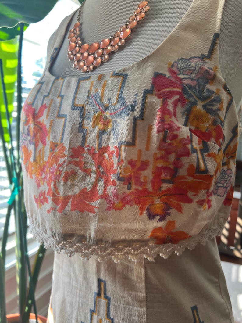 May include: A beige sundress with a floral print in shades of orange, pink, and blue. The dress features a scoop neckline, a decorative lace trim, and a geometric pattern. A peach-colored necklace is draped over the dress.