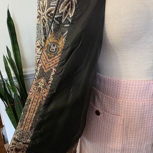 May include: A patterned jacket with a black interior lining. The jacket features a detailed design with brown, gold, and cream colors. The lining is black. The jacket is paired with a pink skirt.