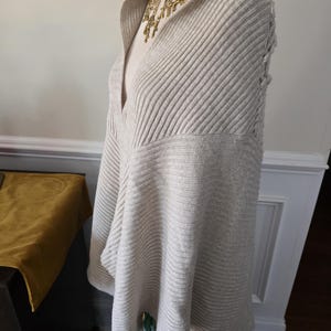 May include: A light beige, ribbed knit poncho with a draped cowl neck. The poncho is displayed on a mannequin and paired with a gold-tone statement necklace. The bottom of the mannequin is wearing a colourful floral skirt.