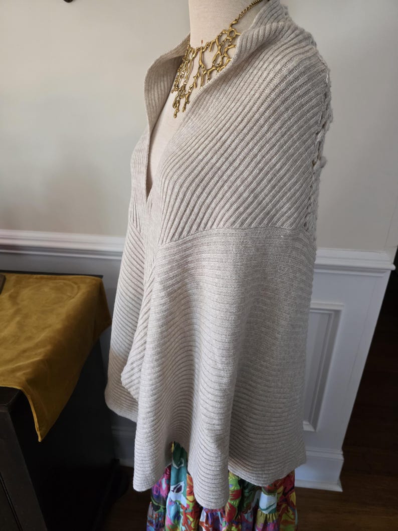 May include: A light beige, ribbed knit poncho with a draped cowl neck. The poncho is displayed on a mannequin and paired with a gold-tone statement necklace. The bottom of the mannequin is wearing a colourful floral skirt.