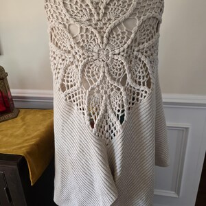 May include: A cream-coloured, open-front cardigan with a large, intricate crochet back panel. The back features a floral design with openwork details. The cardigan has ribbed knit sleeves and a draped front, offering a cosy and stylish look.