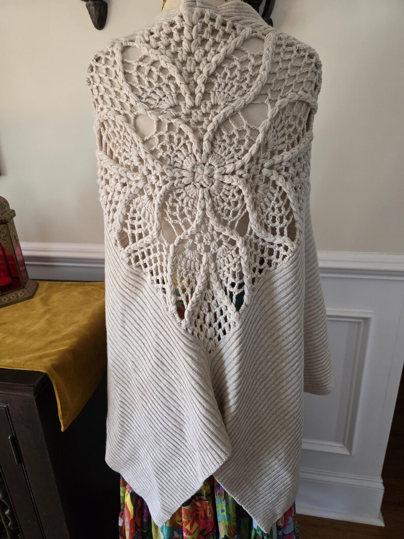 May include: A cream-coloured, open-front cardigan with a large, intricate crochet back panel. The back features a floral design with openwork details. The cardigan has ribbed knit sleeves and a draped front, offering a cosy and stylish look.