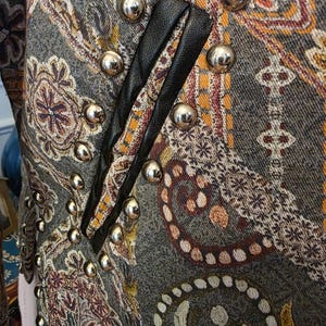 May include: Close-up of a patterned jacket with silver metal buttons. The jacket features a paisley design in shades of gray, brown, and orange. Black leather trim accents the front. The jacket has a vintage or bohemian aesthetic.