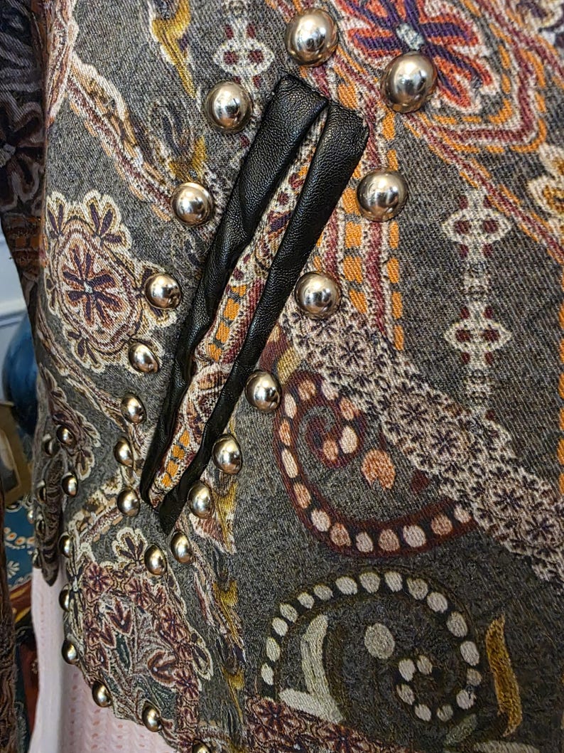 May include: Close-up of a patterned jacket with silver metal buttons. The jacket features a paisley design in shades of gray, brown, and orange. Black leather trim accents the front. The jacket has a vintage or bohemian aesthetic.