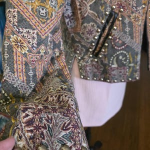 May include: Close-up of a patterned jacket with a mix of colors including gray, gold, and burgundy. The jacket features a detailed floral and paisley design, with gold-colored studs along the edges and on the sleeves. The jacket has a black pocket.