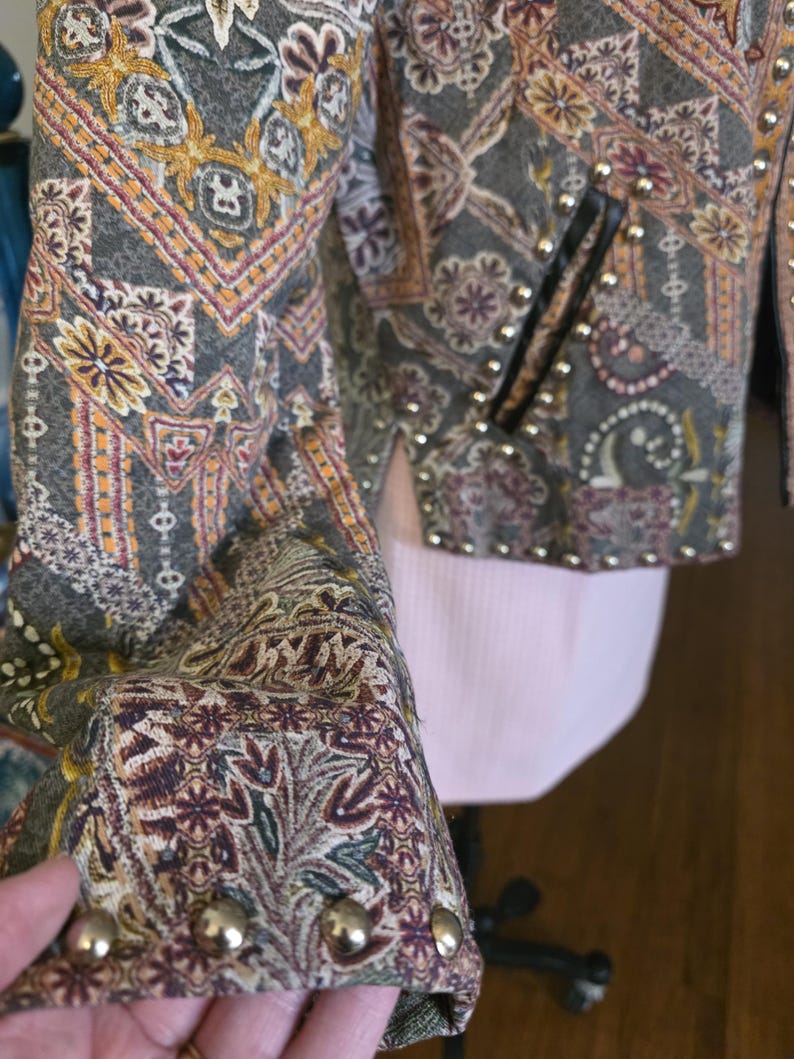 May include: Close-up of a patterned jacket with a mix of colors including gray, gold, and burgundy. The jacket features a detailed floral and paisley design, with gold-colored studs along the edges and on the sleeves. The jacket has a black pocket.