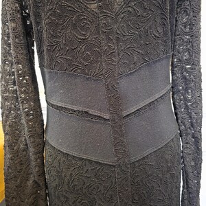 May include: A black lace dress with long sleeves and a fitted waist. The dress features a floral pattern and a contrasting solid black band at the waist. The sleeves are also made of lace.