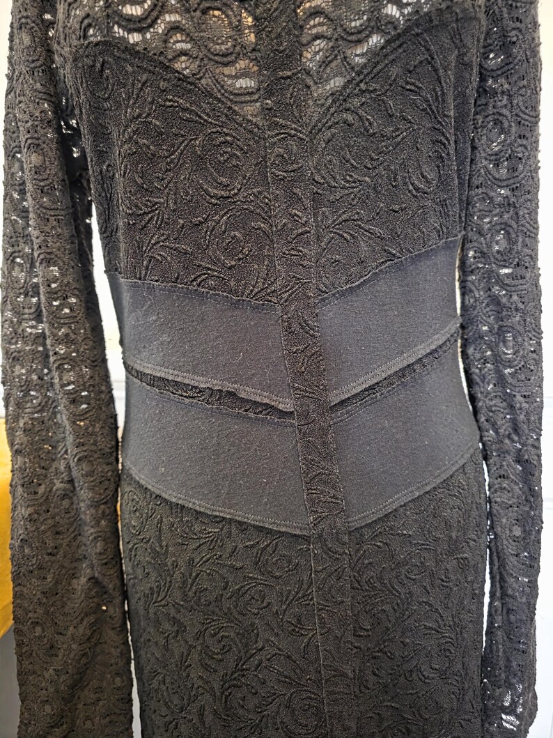 May include: A black lace dress with long sleeves and a fitted waist. The dress features a floral pattern and a contrasting solid black band at the waist. The sleeves are also made of lace.