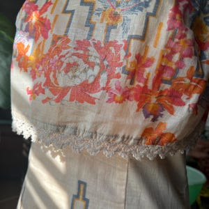 May include: A beige dress with a floral and geometric pattern. The top section features a vibrant floral design in shades of orange, pink, and yellow, with a geometric pattern in blue and yellow. The dress has a lace trim along the neckline.