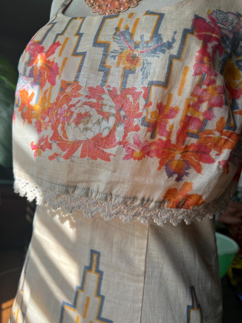 May include: A beige dress with a floral and geometric pattern. The top section features a vibrant floral design in shades of orange, pink, and yellow, with a geometric pattern in blue and yellow. The dress has a lace trim along the neckline.
