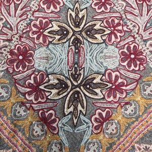 May include: Close-up of a textile with an intricate floral pattern. The design features burgundy, gold, and light blue embroidered flowers and geometric shapes on a gray background. The overall aesthetic is ornate and detailed.
