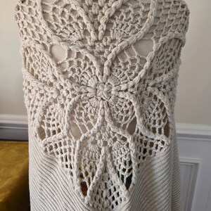 May include: A cream-coloured, open-front cardigan with a detailed crochet back panel. The back features a large, floral-like design with intricate openwork and braided edging. The body of the cardigan has a ribbed knit texture.