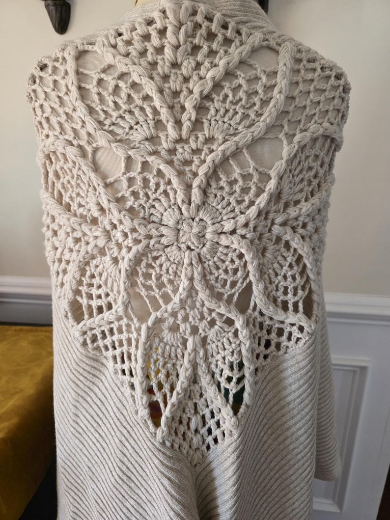 May include: A cream-coloured, open-front cardigan with a detailed crochet back panel. The back features a large, floral-like design with intricate openwork and braided edging. The body of the cardigan has a ribbed knit texture.