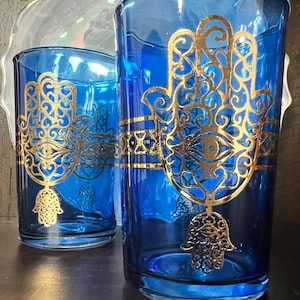 May include: Two blue drinking glasses with gold Hamsa hand designs. The glasses are different sizes and are decorated with intricate gold detailing. The background includes a decorative, iridescent plate.
